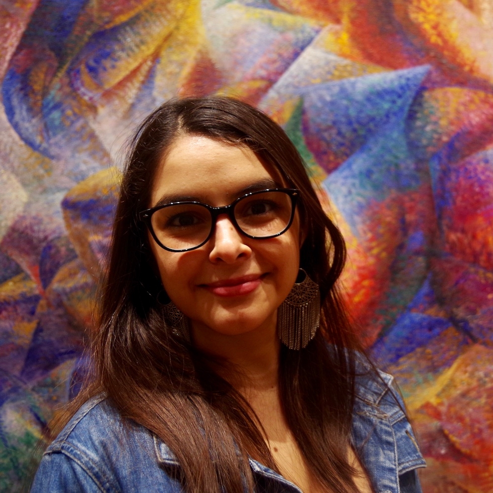 Maria Belen Contreras headshot. Long brown hair, glasses, standing in front of blue and red wall wearing denim shirt